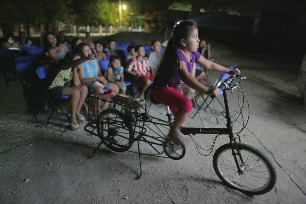 A girl pedals to generate electricity for a movie projector as her family watches